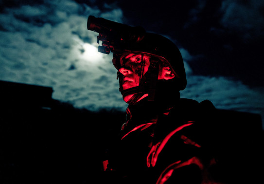 A soldier glows red while standing in front of moon-lit clouds wearing a helmet and Army uniform at night.