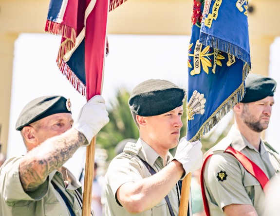 Three army personnel stand holding colourful flags whilst on a parade.
