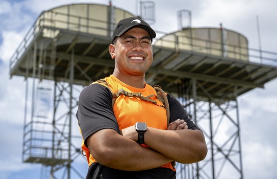 A man in an orange high vis t-shirt with black sleeves and a black baseball cap, smiles at the camera with his arms crossed with a large metal structure in the background.